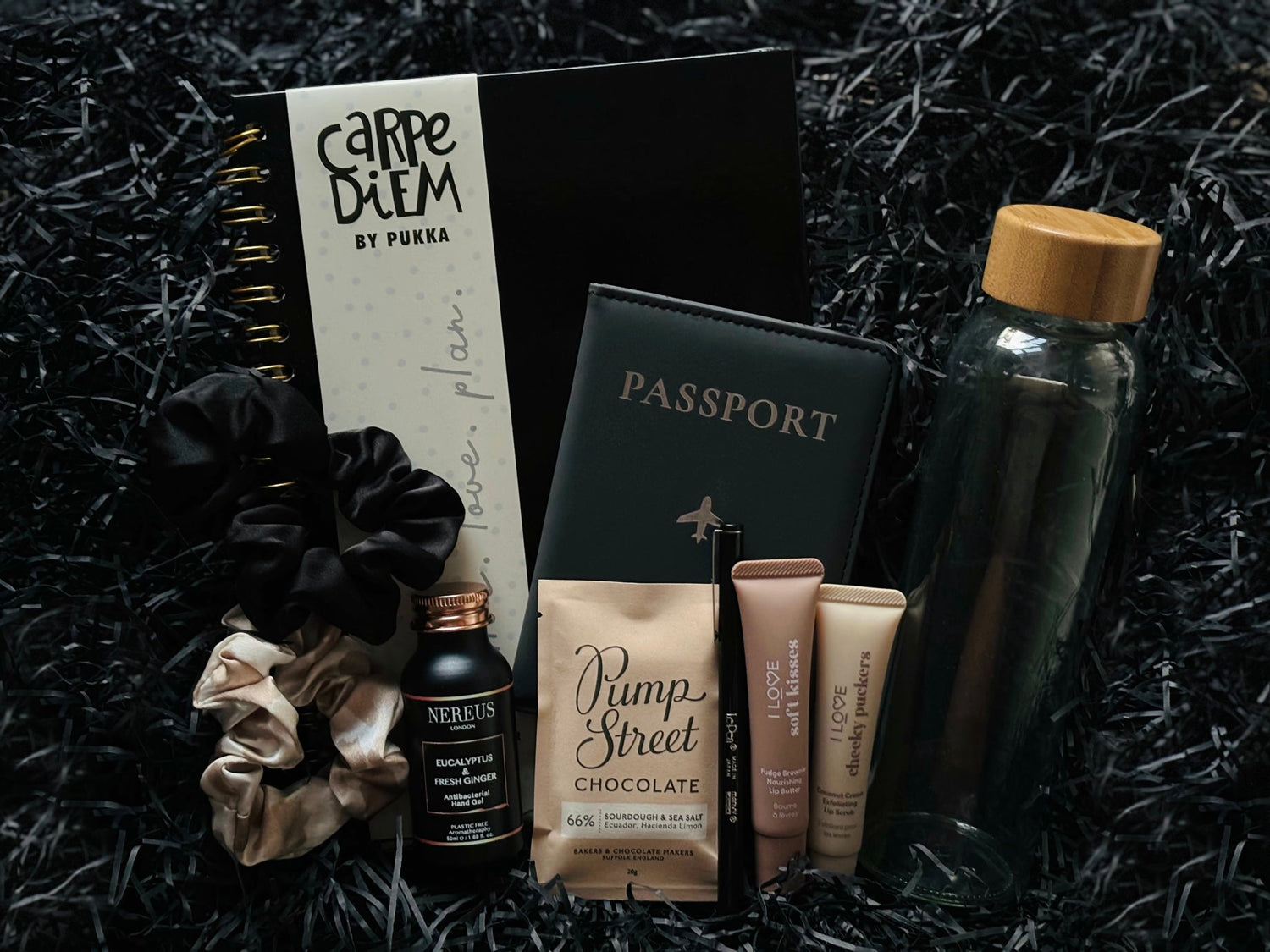 Products, L to R: two Kitsch scrunchies, Nereus hand gel, a pukka notebook, a passport cover, , Pump Street Chocolate, a Marvy Uchida fineliner pen, I Love Cosmetics lip butter and lip scrub, and a glass water bottle.