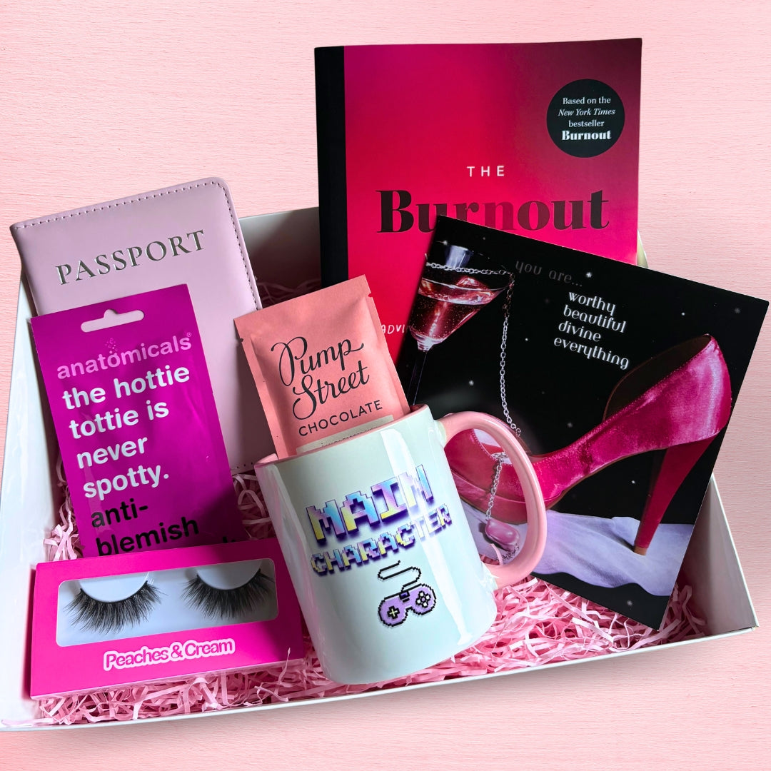 A pink themed gift box. Contents: false eyelashes, a face mask, a passport cover, chocolate, a mug, a book, and a greeting card.
